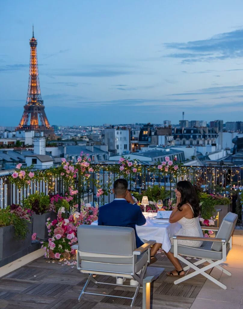 Couple dining at a private rooftop table with Eiffel Tower views at dusk, surrounded by pink floral arrangements and string lights, Paris