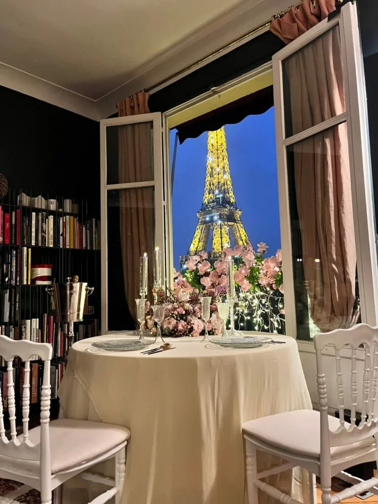 Intimate private dinner table set for two in a Parisian apartment, with candlelight, pink flowers, and a direct view of the illuminated Eiffel Tower through an open window