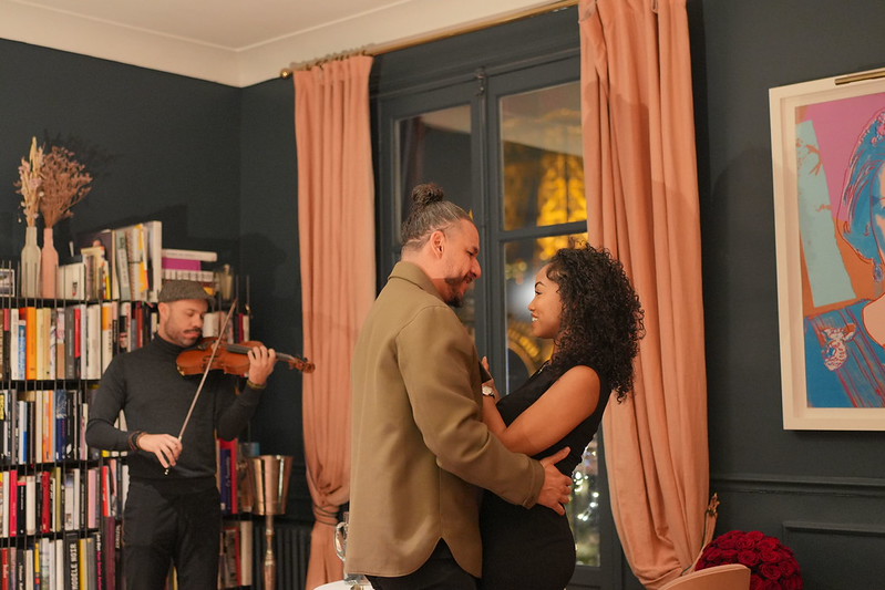 Couple sharing an intimate moment during a private dinner in a Parisian apartment, with a live violinist performing in the background and the Eiffel Tower visible through the window