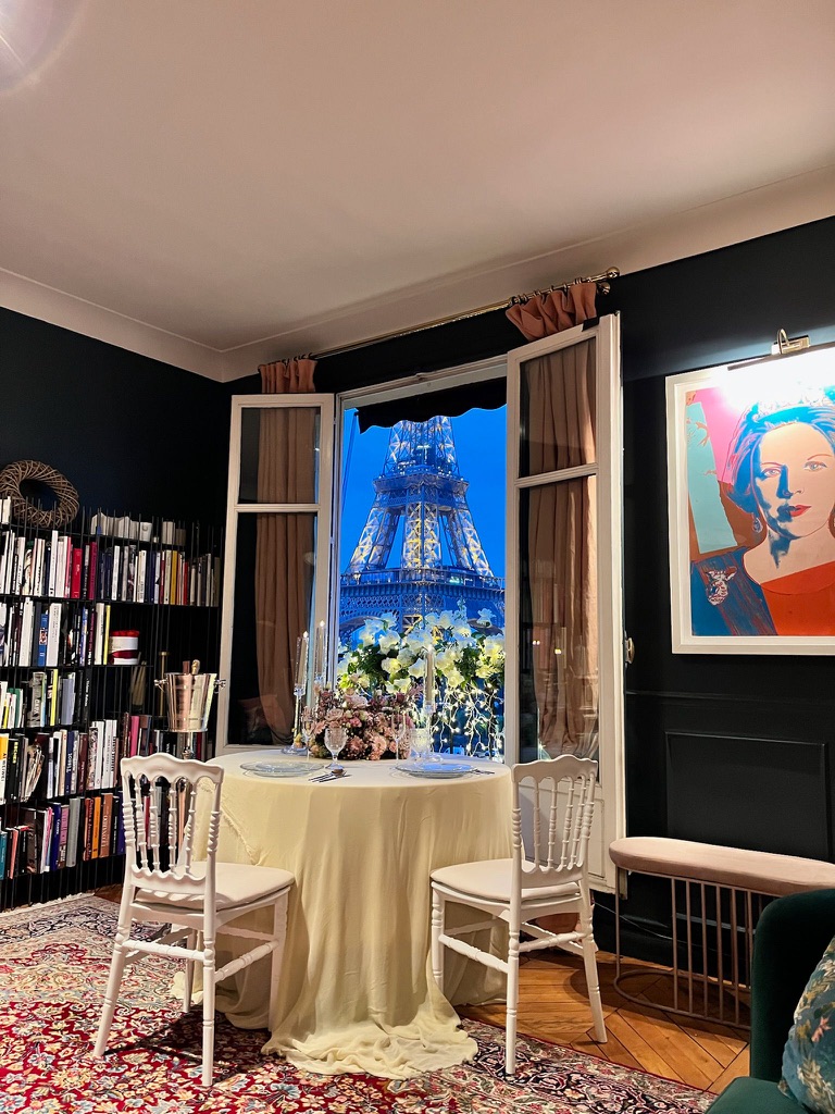Private dinner table for two set in a Parisian apartment on Avenue de New York, with candlelight, flowers, and a direct view of the illuminated Eiffel Tower through open French windows