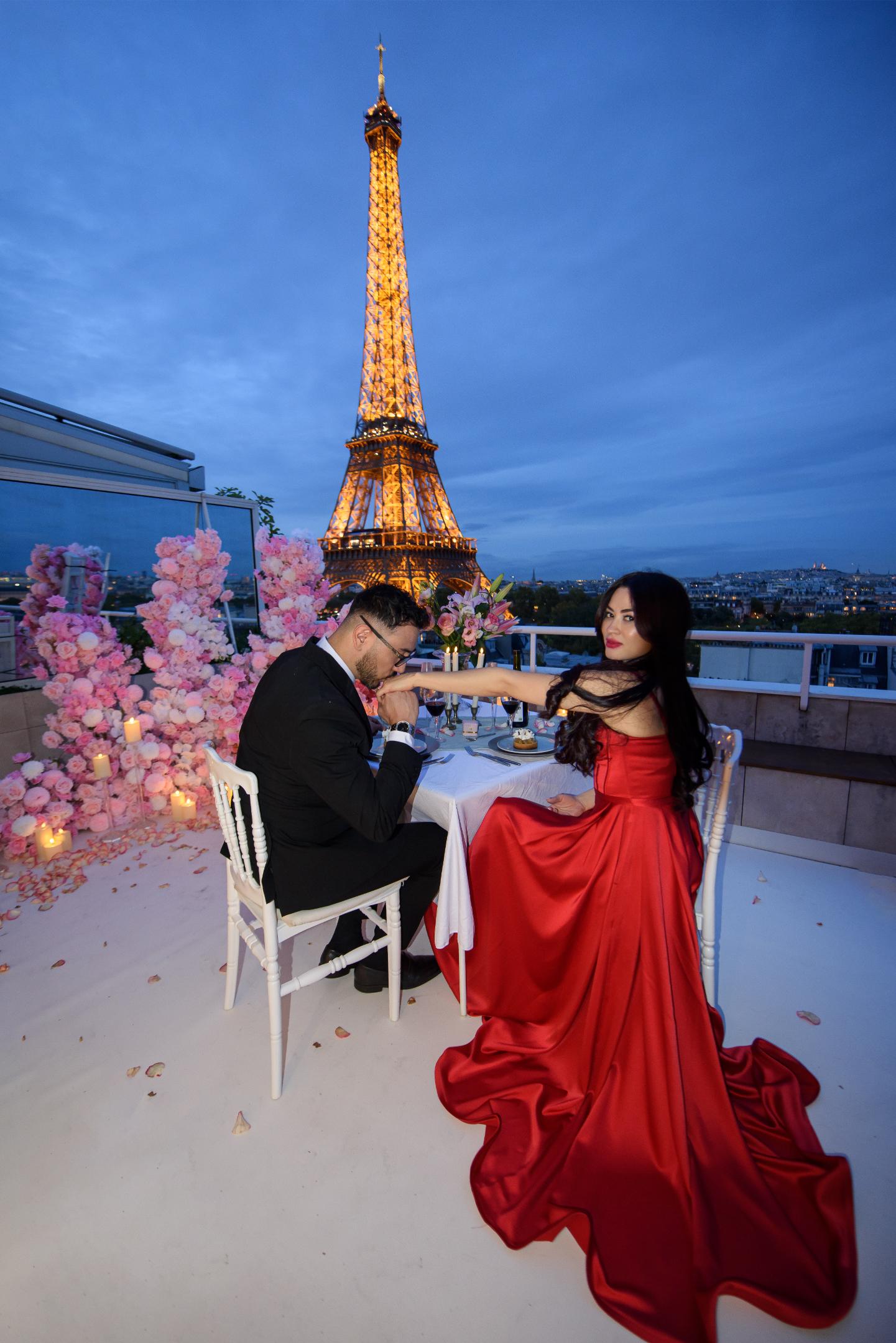 Couple at a private rooftop dinner table on La Terrasse Eiffel in Paris, with pink floral arrangements, candles, and the illuminated Eiffel Tower directly behind them at dusk