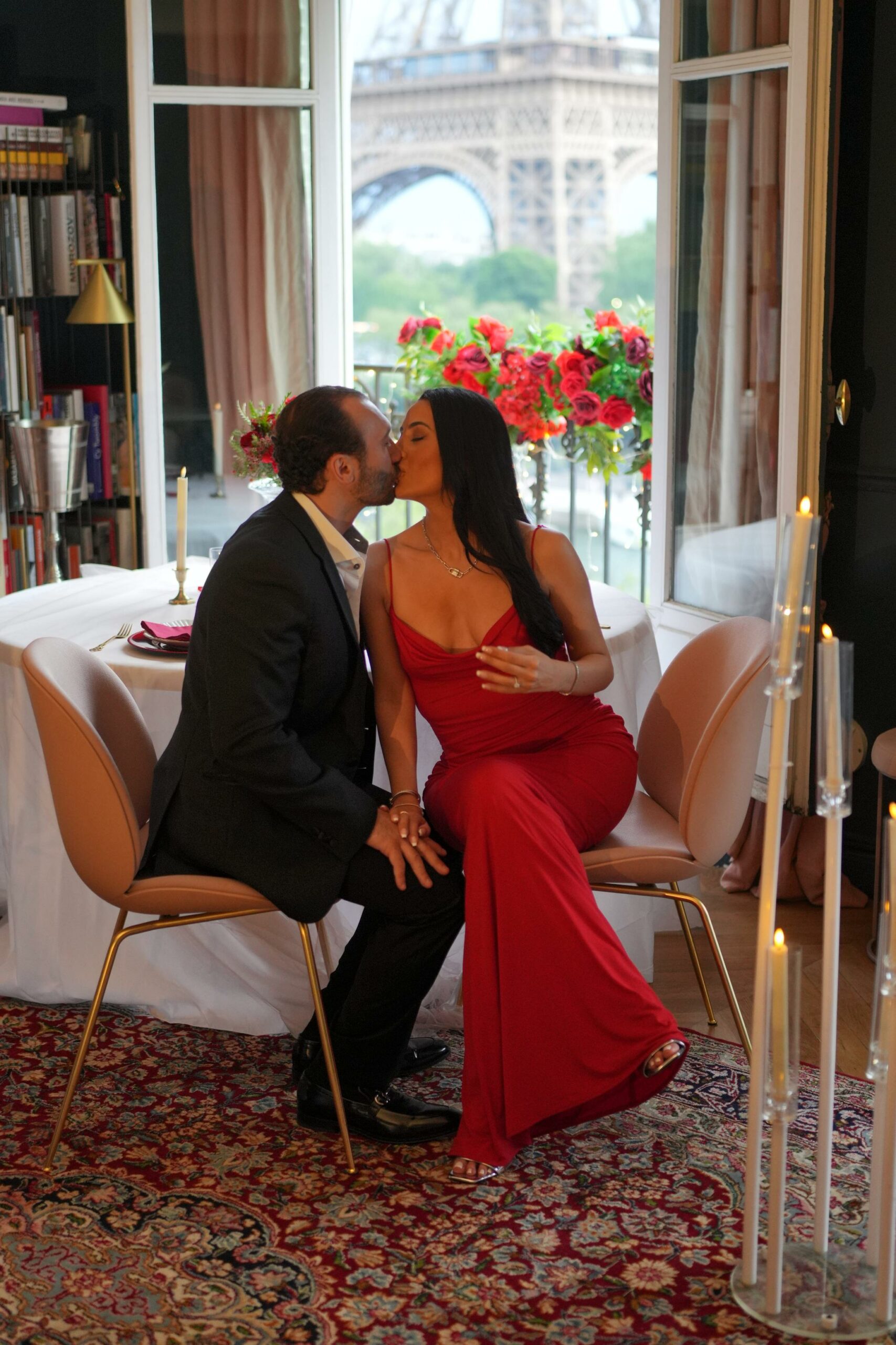 Couple sharing a kiss at a private dinner table in a Parisian apartment, with red roses and the Eiffel Tower visible through the open French windows