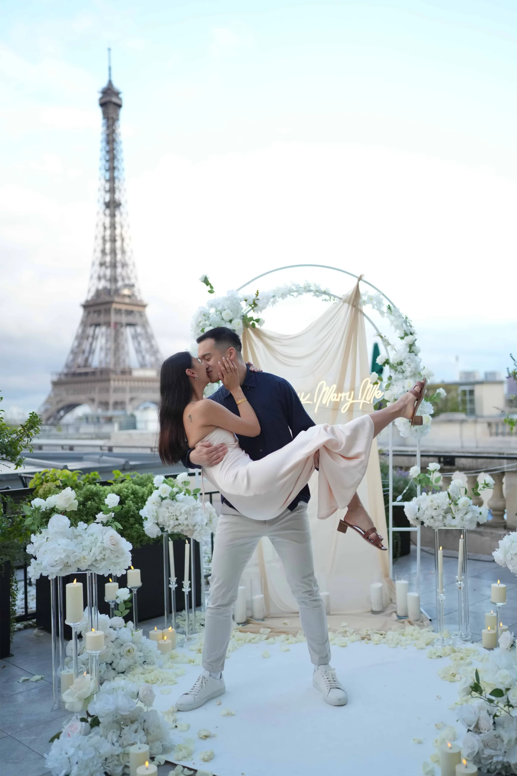 Couple s'embrassant sur un rooftop privatisé avec décoration florale blanche, arche et néon "Marry Me" face à la Tour Eiffel à Paris