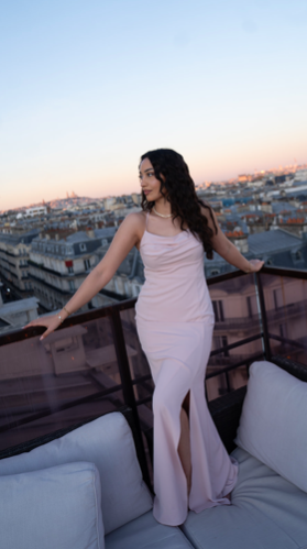 Woman in a long blush pink dress standing on a Paris rooftop terrace at sunset, with Parisian rooftops behind her, dressed up for a proposal