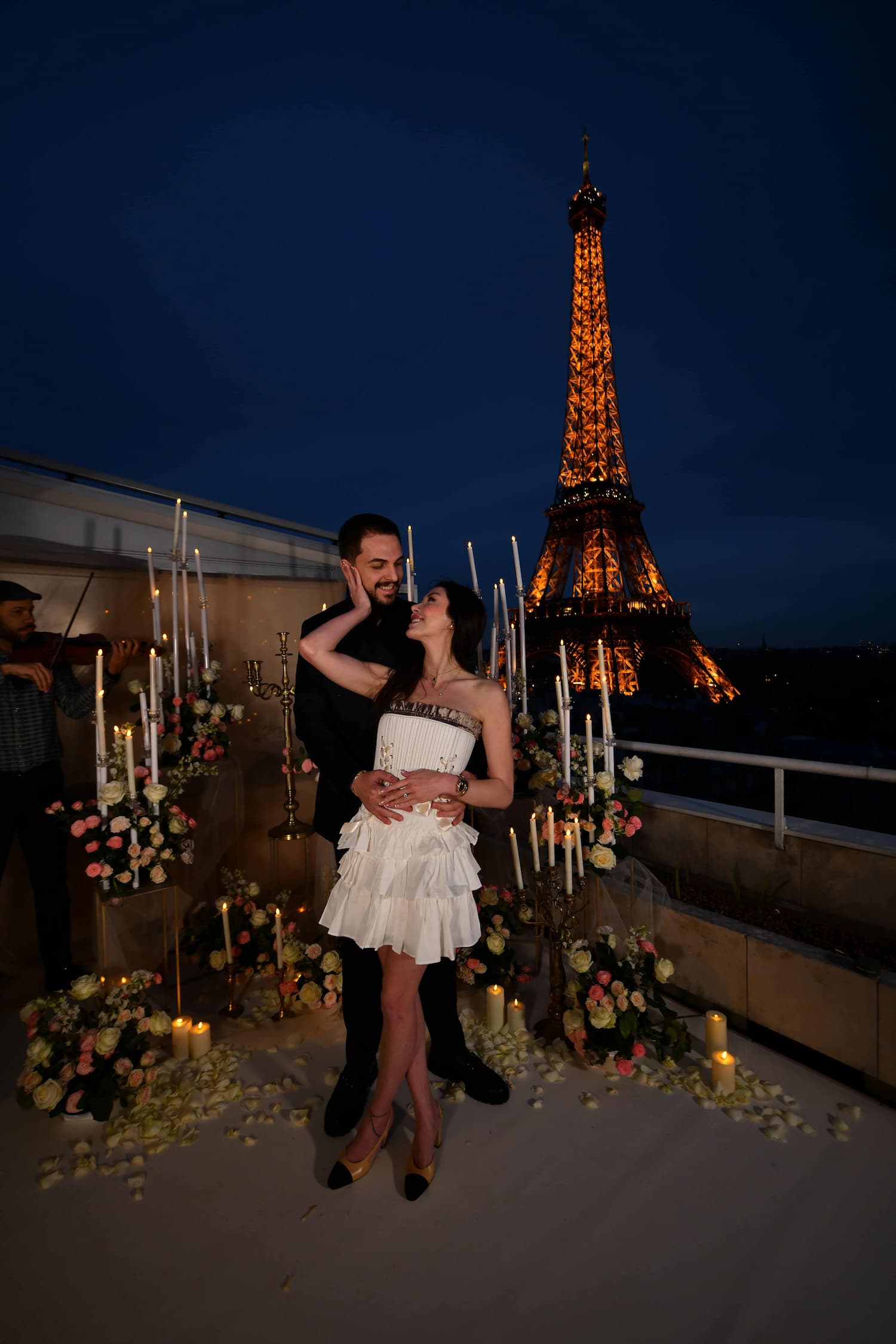 Newly engaged couple embracing on the Terrasse Eiffel private rooftop in Paris after the proposal, surrounded by candles and pale roses with a live violinist and the illuminated Eiffel Tower