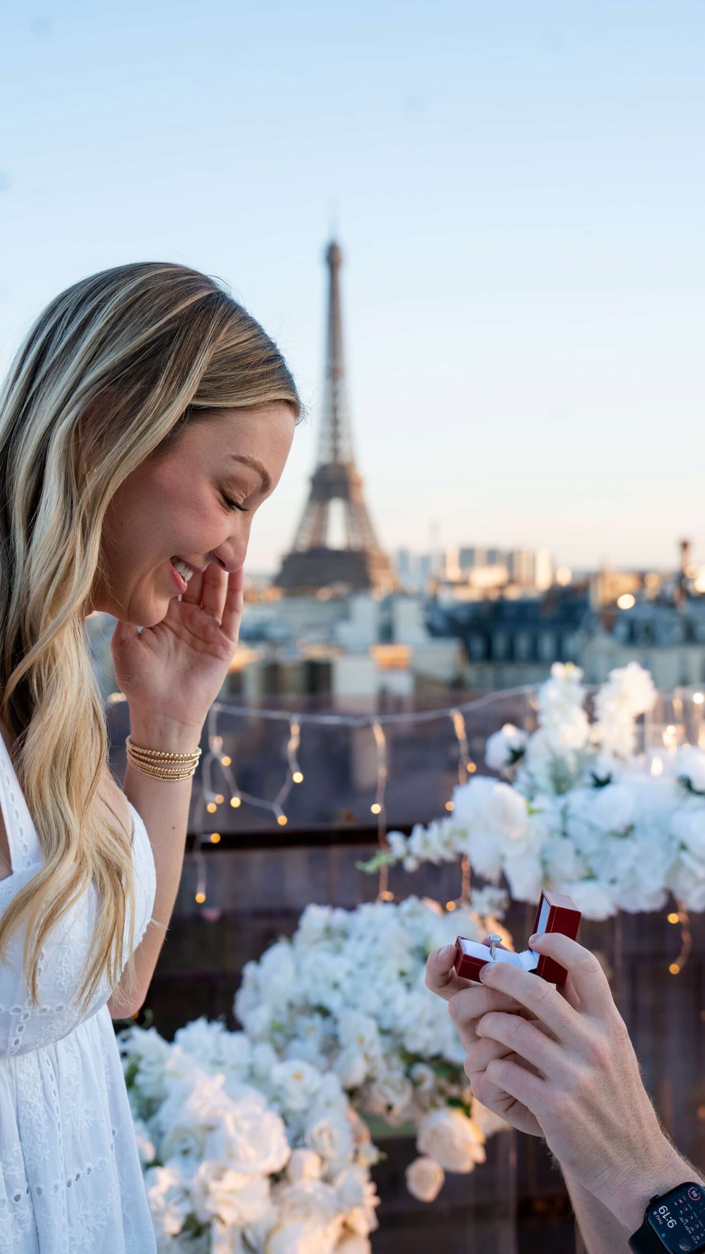Woman covering her mouth in surprise as her partner opens a red ring box on a Paris terrace, with white flowers, fairy lights and the Eiffel Tower in the background