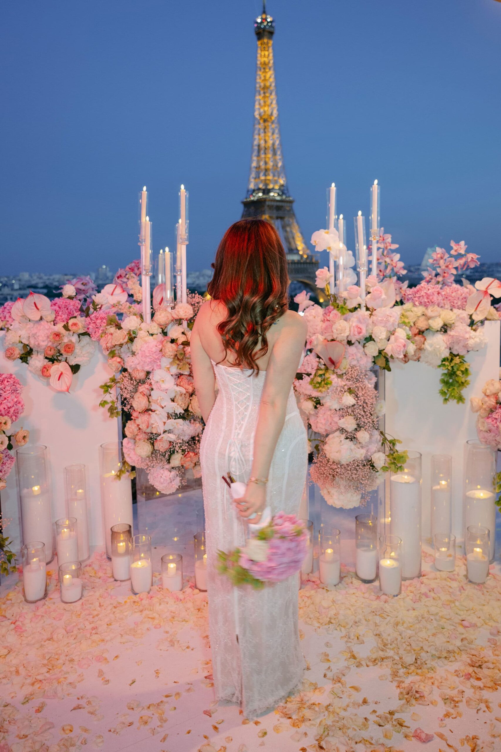 Woman in a white dress seen from behind discovering a pink and white floral proposal setup with candles and rose petals on a Paris rooftop at blue hour, facing the illuminated Eiffel Tower