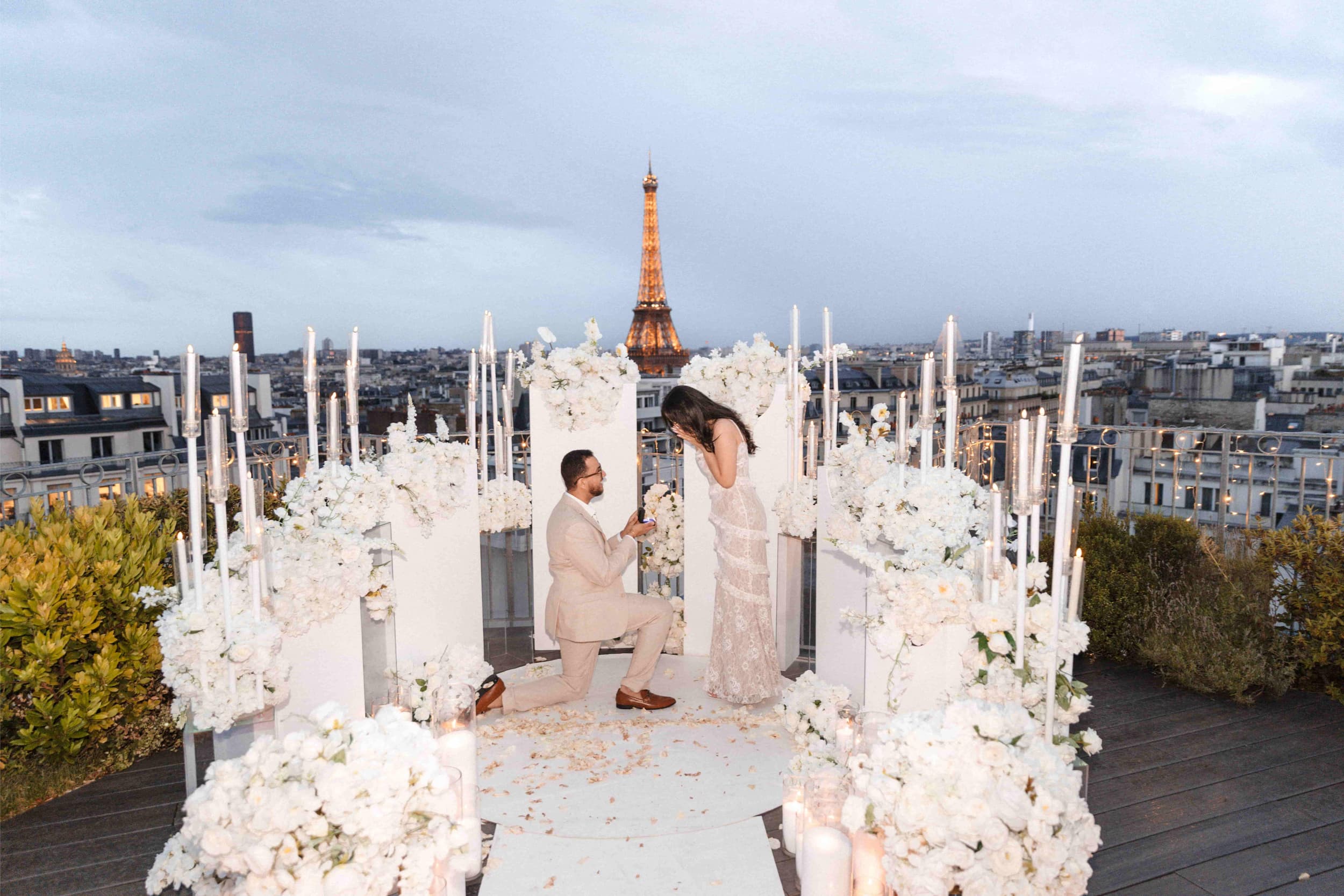 Man proposing on one knee on a private rooftop in Paris at blue hour, surrounded by white flowers and candles with the illuminated Eiffel Tower in the background