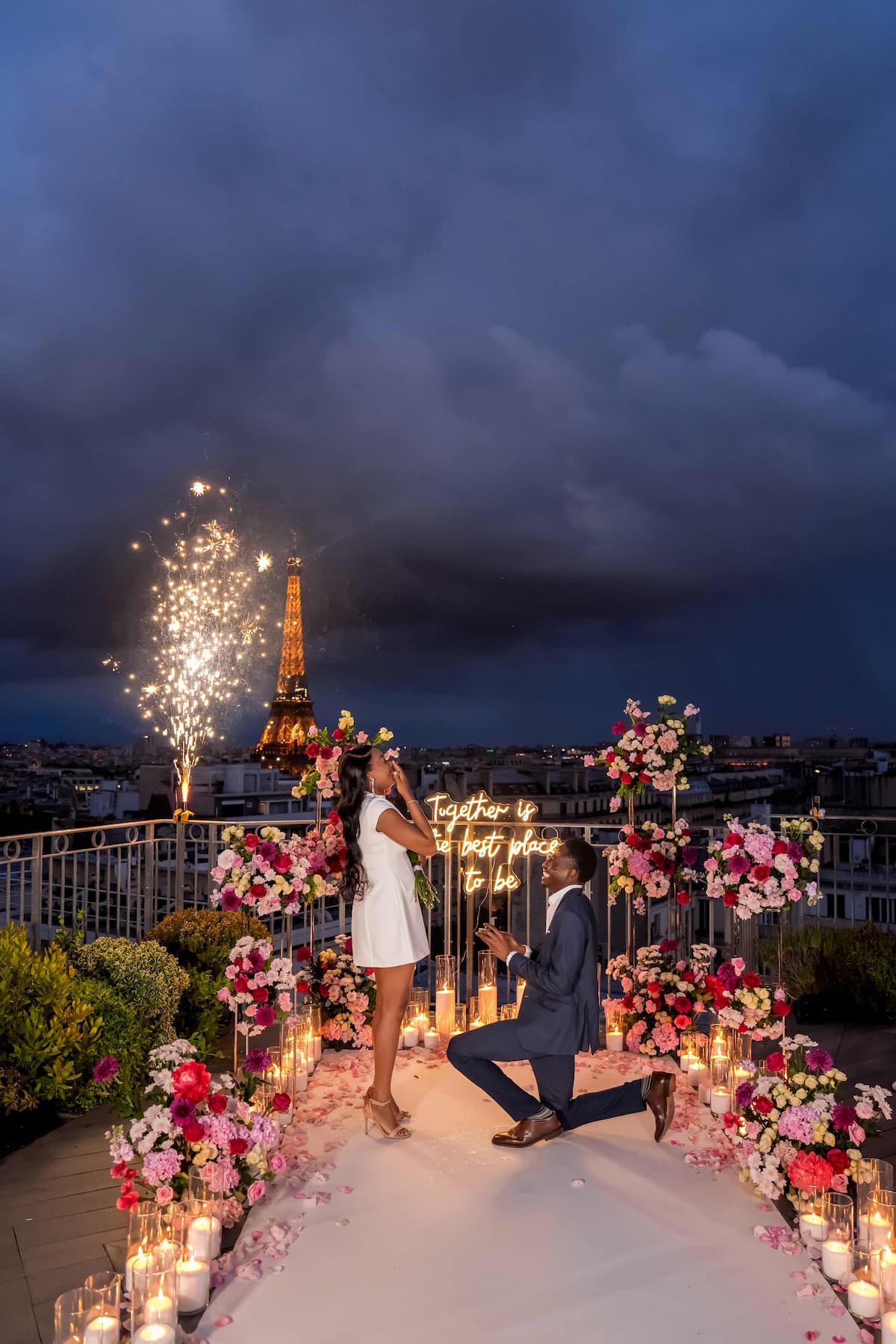 Man kneeling to propose on a private Paris rooftop at night, with pink and red floral arrangements, candles, a cold firework and a "Together is the best place to be" neon sign facing the illuminated Eiffel Tower