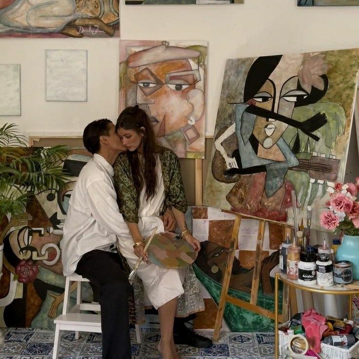 A couple sharing a tender kiss in an artist's studio, surrounded by expressive paintings and art supplies.