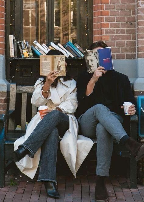 A couple sitting on a bench outside a bookshop, each hiding their face behind a book of memories while holding a coffee cup