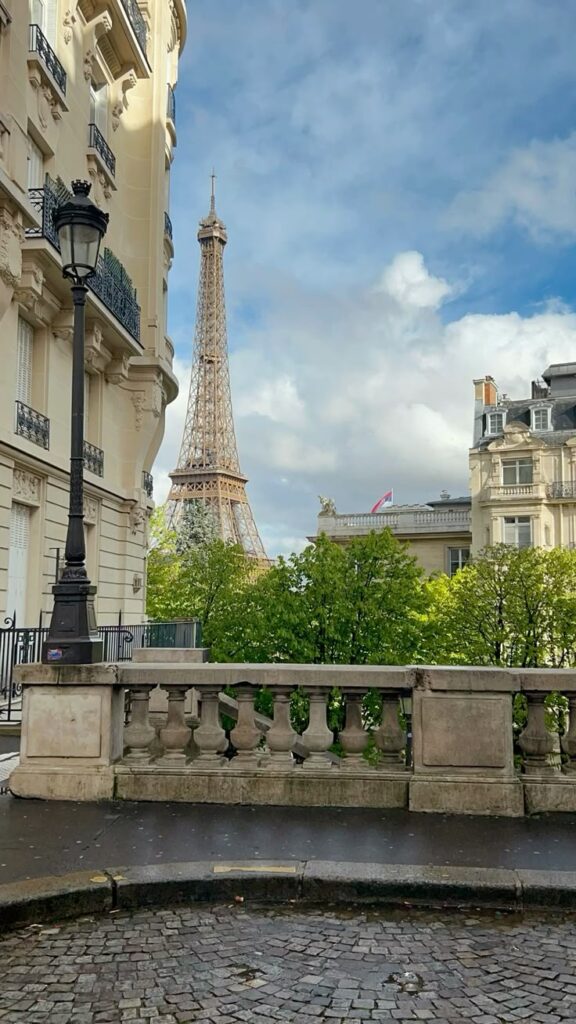 Alt text:
"View of the Eiffel Tower rising above lush green trees on Avenue de Camoëns in Paris, framed by a classic stone balustrade, a vintage black street lamp, and elegant Haussmann apartment buildings on either side under a dramatic cloudy sky