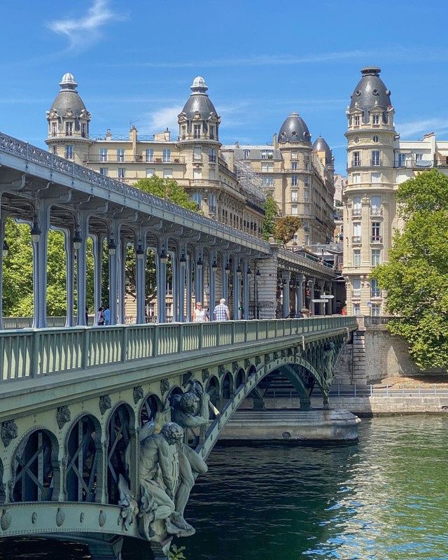 Close-up view of the Bir-Hakeim bridge in Paris on a sunny day, featuring its ornate green iron structure with decorative columns and arches, a sculpted stone statue group in the foreground, the Seine river below, and elegant Haussmann buildings with grey mansard rooftops in the background