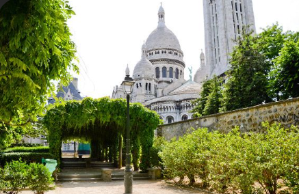 A sunlit pathway through the Parc de Bagatelle rose garden in Paris, framed by black iron arches covered in blooming pink climbing roses, leading through a series of gates flanked by neatly trimmed box hedges, with visitors relaxing on the grass under a bright blue sky