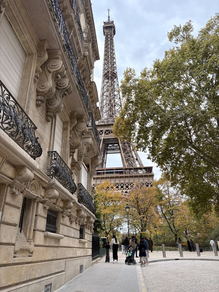 Street-level view of the Eiffel Tower rising above Haussmann-style buildings with ornate stone facades and wrought iron balconies on Rue de l'Université in Paris, framed by autumn trees with golden leaves, with a few pedestrians walking along the cobblestone pavement below