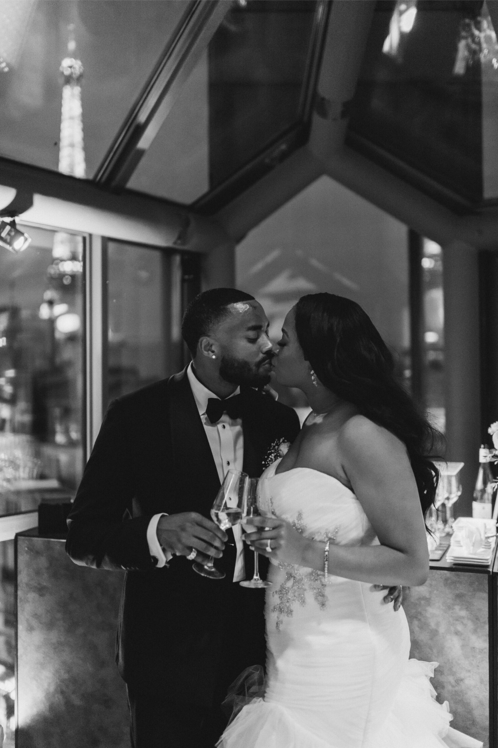Newlywed couple sharing a champagne kiss during their elopement dinner in Paris, Eiffel Tower visible through the window