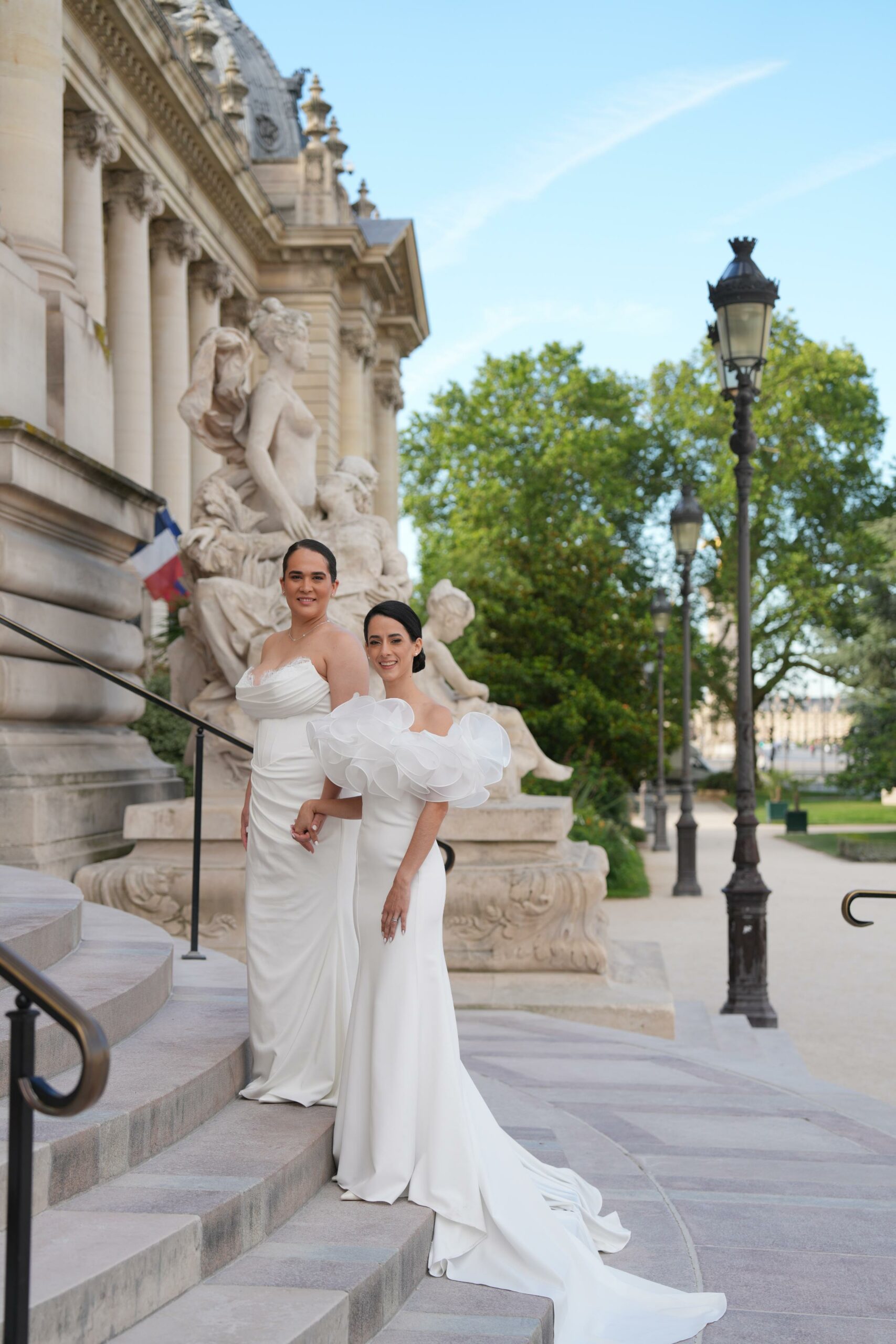 Two brides in elegant white wedding gowns posing on the steps of the Petit Palais during their Paris elopement