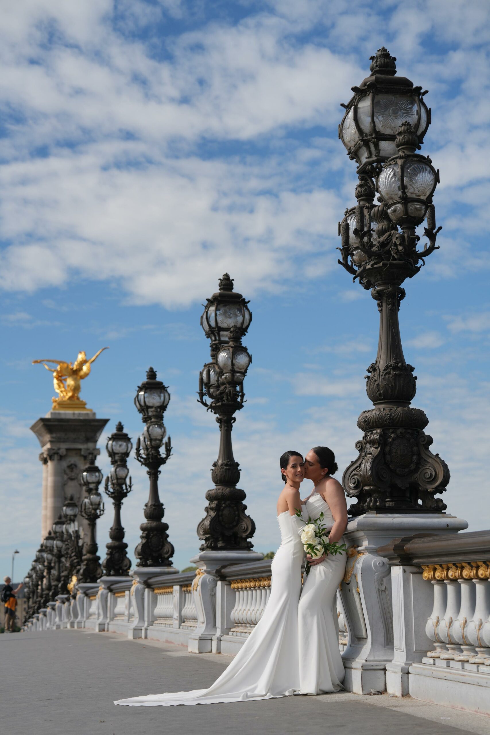 Two brides sharing a kiss on Pont Alexandre III during their Paris elopement, ornate lampposts and golden statue in the background