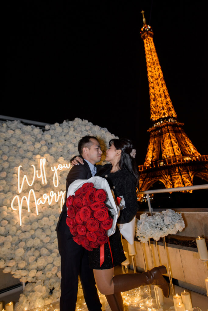 A man offering a large bouquet of red roses to his partner on a romantic rooftop terrace, with the illuminated Eiffel Tower glowing in the night sky behind them.