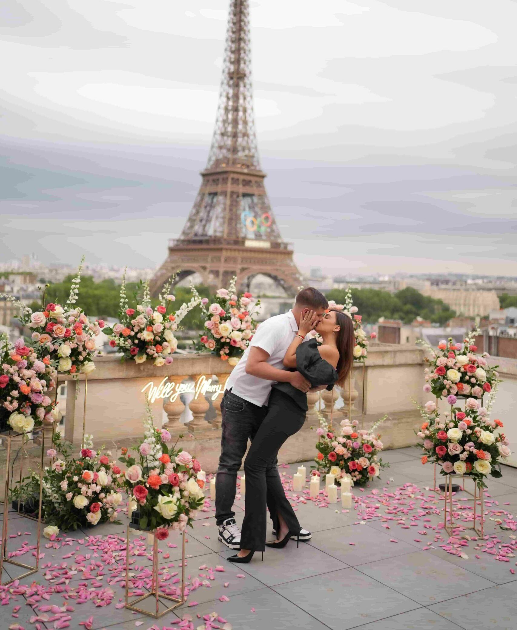 A couple sharing a kiss during a marriage proposal on a rooftop terrace in Paris, surrounded by lush floral arrangements in pink, coral and white, with the Eiffel Tower in the background and a neon 'Will you marry me?' sign