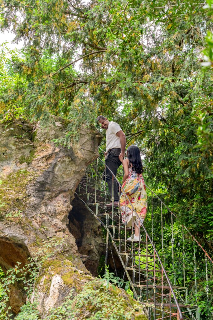 A couple climbing a rustic iron staircase carved into a mossy rock, surrounded by lush greenery, sharing a smile in a secret garden setting.