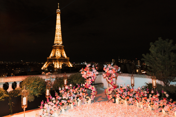 Rooftop decorated with pink and peach floral arrangements facing the illuminated Eiffel Tower at night, with candles and golden lanterns creating a romantic atmosphere.