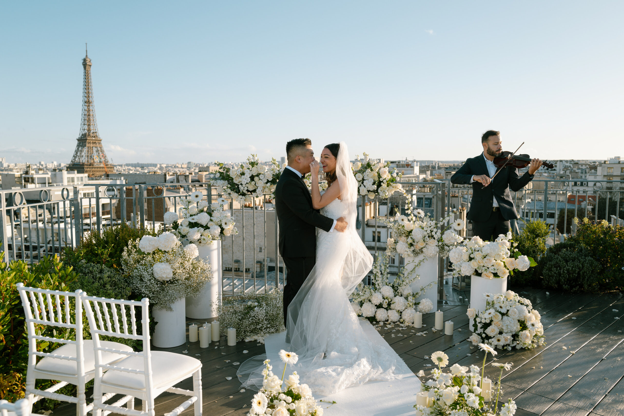 Bride and groom sharing a joyful dance on a rooftop terrace overlooking Paris and the Eiffel Tower, surrounded by white floral arrangements and candles, with a violinist performing beside them.