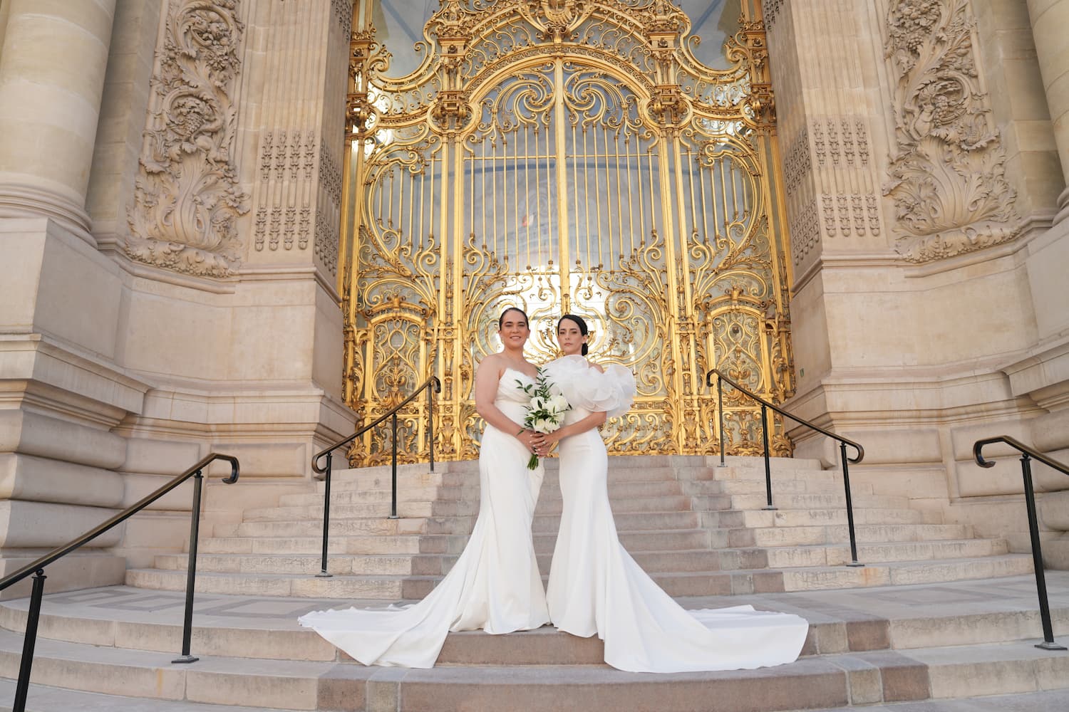Same-sex couple in white wedding gowns standing before the golden gate at Petit Palais during their Paris elopement ceremony