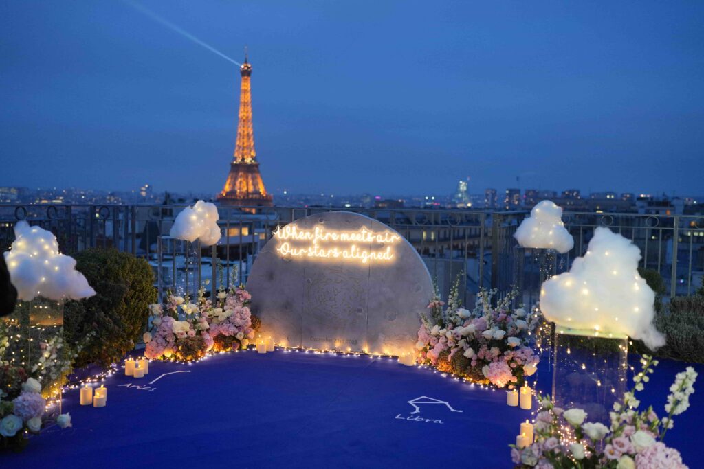 A romantic rooftop proposal setup in Paris with candles, pastel flowers, and a glowing neon sign, with the Eiffel Tower illuminated in the background at night.