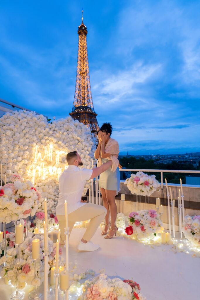 A man proposing on one knee during a romantic rooftop setup filled with flowers and candles, with the Eiffel Tower glowing in the background at dusk.
