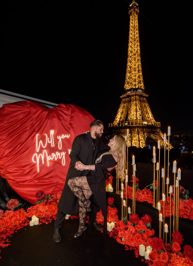 A couple dancing beside a red heart-shaped backdrop with a “Will you marry me” sign, surrounded by candles and red roses, with the Eiffel Tower illuminated at night.