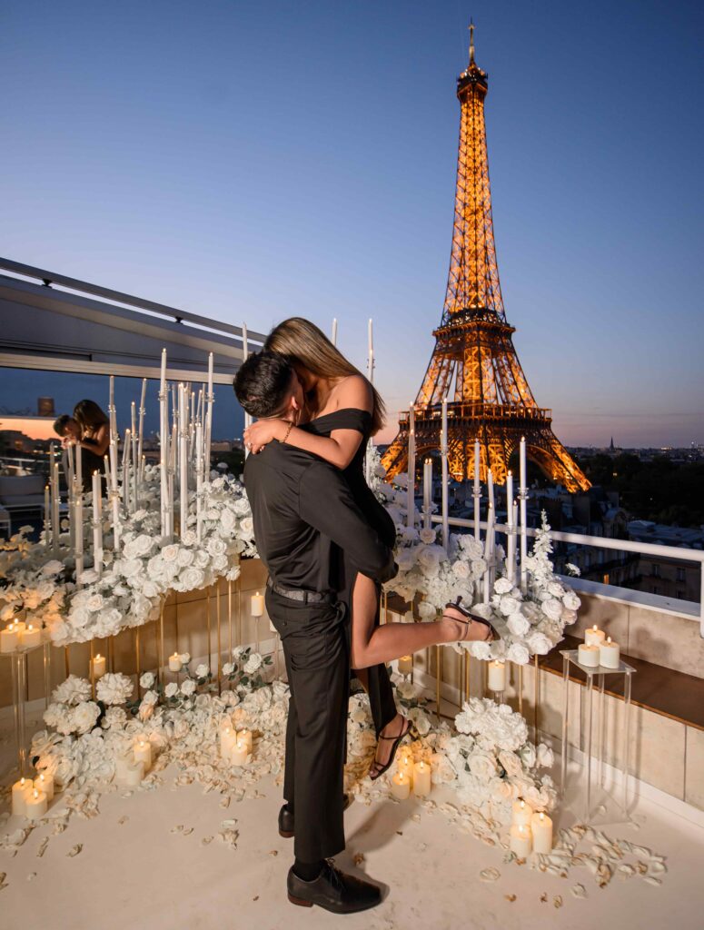 A couple embracing on a flower-filled rooftop terrace with candles, overlooking the illuminated Eiffel Tower at sunset in Paris.
