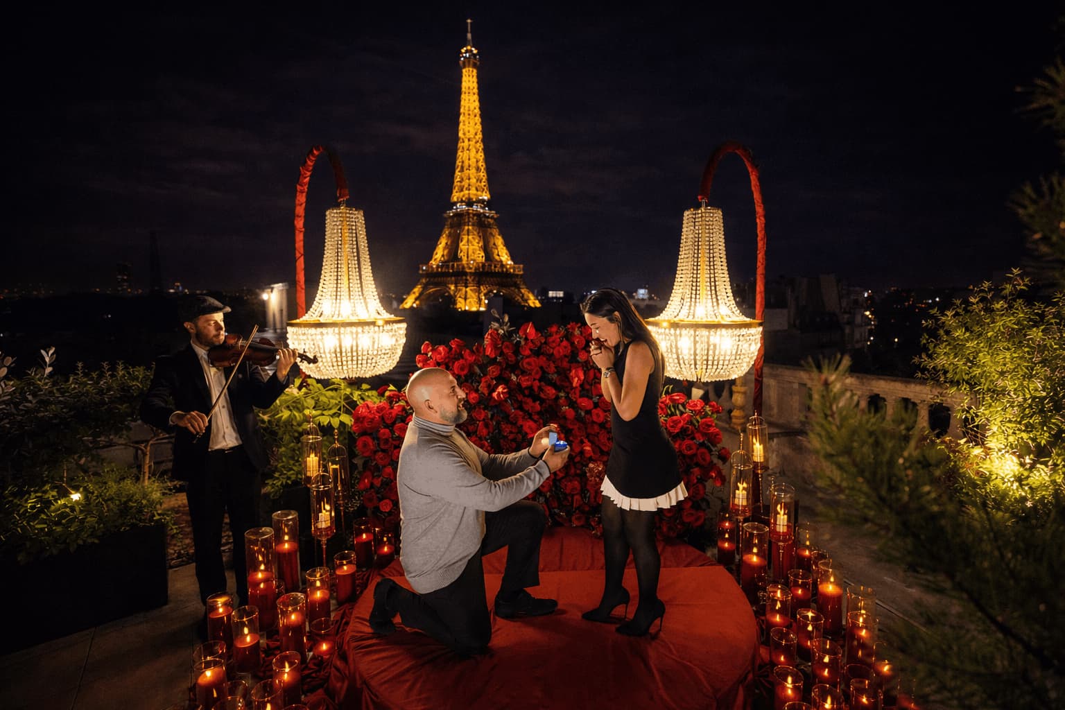 A man proposing on one knee on a Paris rooftop at night, surrounded by red roses, candles, crystal chandeliers, and a violinist, with the Eiffel Tower glowing in the background.