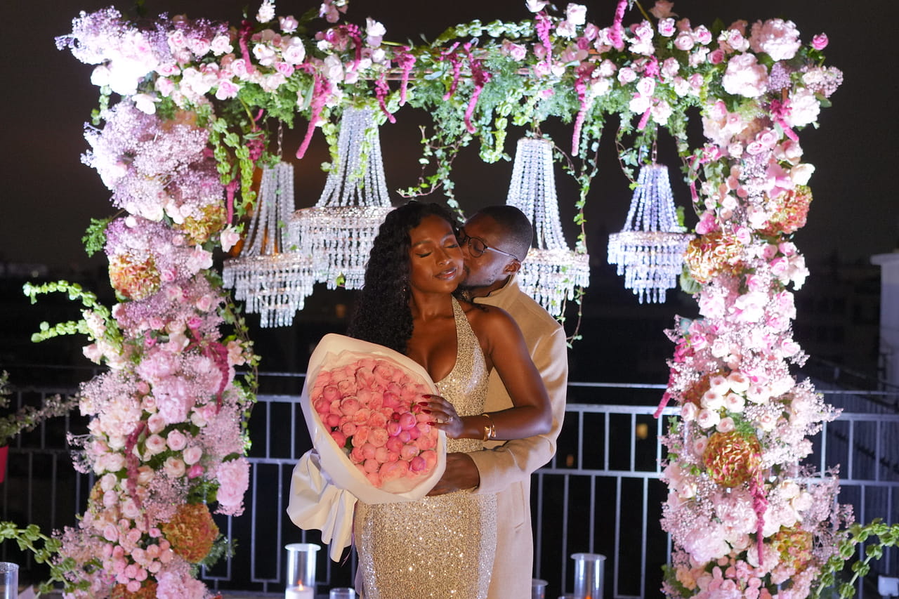 A couple embracing under a luxurious floral arch with crystal chandeliers during a rooftop proposal at night, surrounded by pink flowers and candlelight.