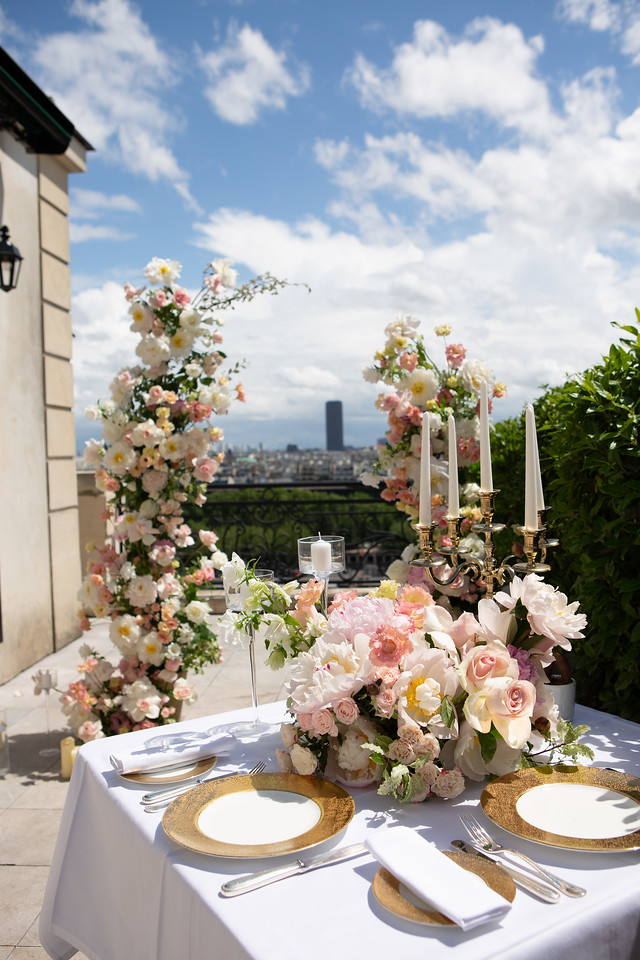 A beautifully decorated romantic table on a Paris terrace with pastel flowers, candles, and a floral arch, overlooking the city under a blue sky.