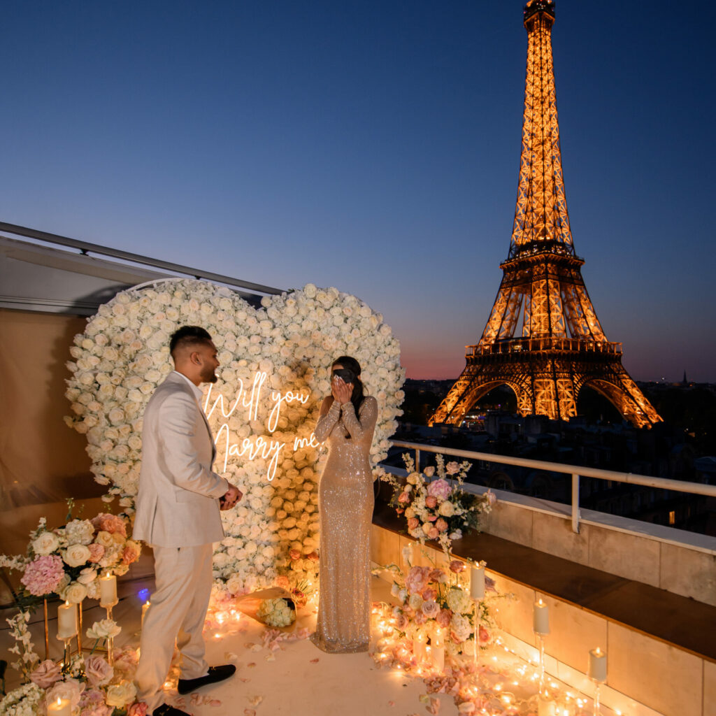 Une femme a les yeux bandés sur un rooftop parisien, avec la Tour Eiffel qui s'illumine en fond. Un décor de fleurs blanches et roses et un coeur blanc avec un néon.