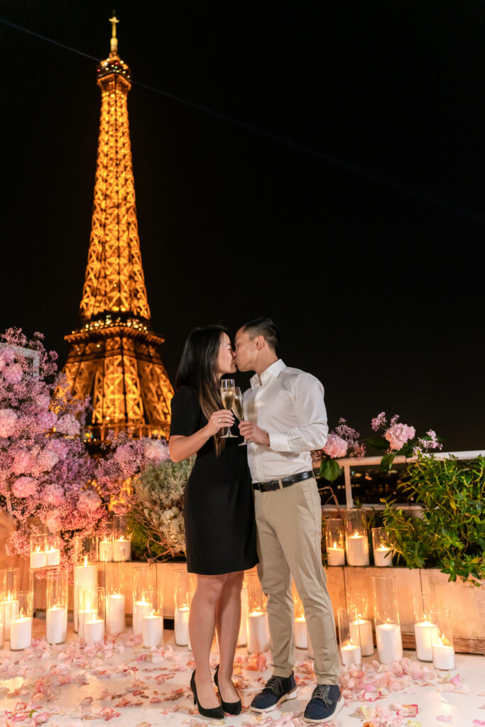 A couple sharing a kiss while holding champagne glasses on a candlelit rooftop, with pink flowers and the illuminated Eiffel Tower behind them at night.