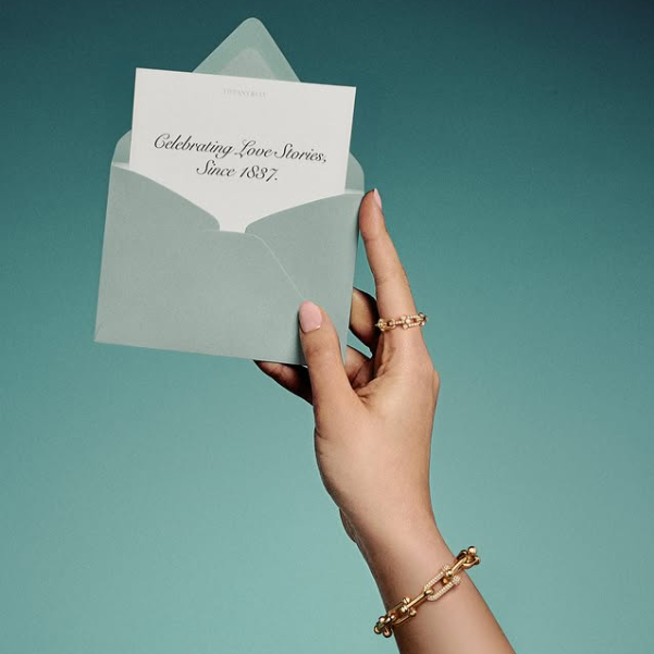 A hand adorned with a Tiffany & Co. gold ring and bracelet holding an open envelope with a card reading 'Celebrating Love Stories Since 1837', symbolising the brand's legacy in engagement jewellery available in Paris.