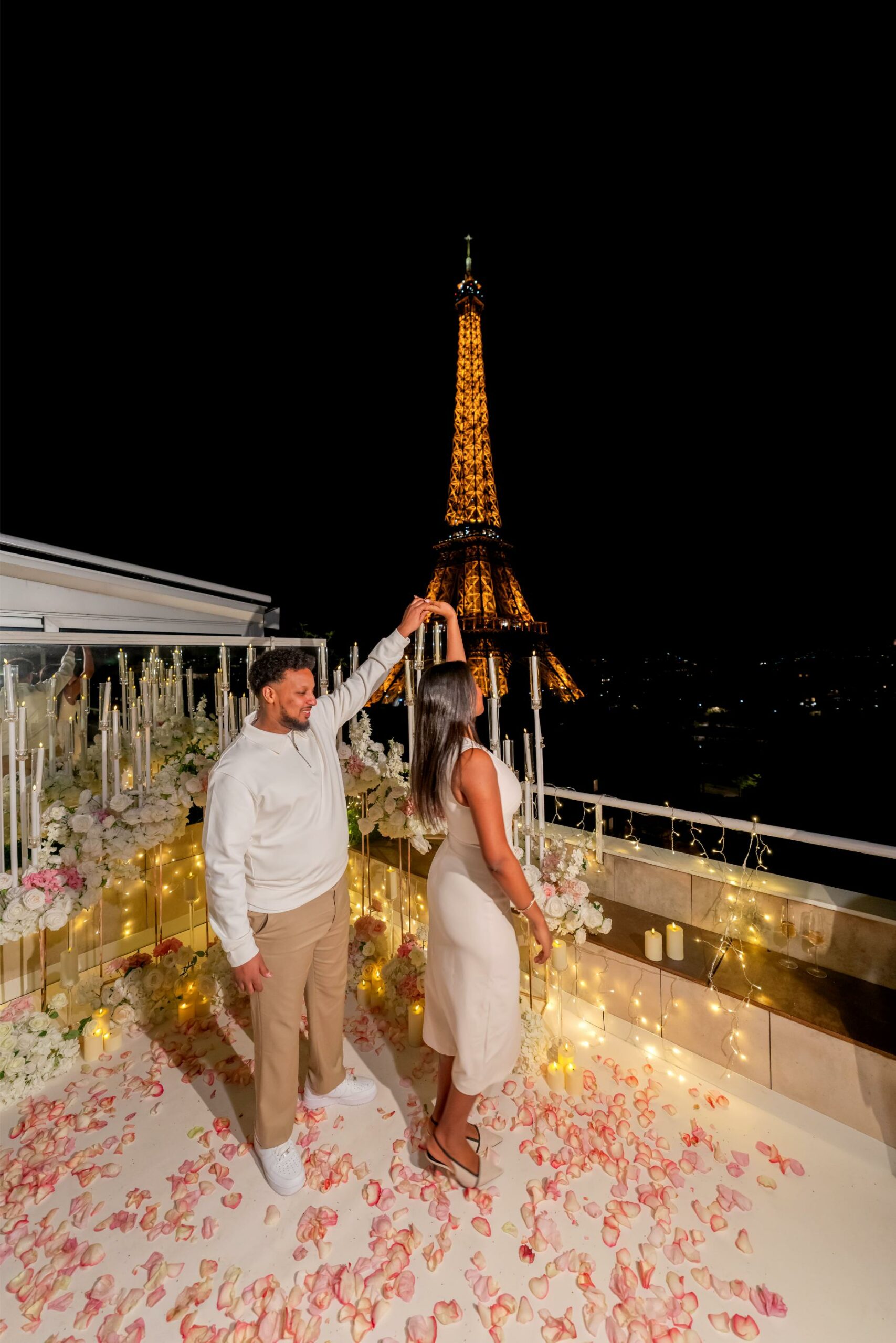 Newly engaged couple dancing on the Eiffel Terrace rooftop at night, surrounded by white flowers, rose petals and fairy lights, with the illuminated Eiffel Tower directly behind them