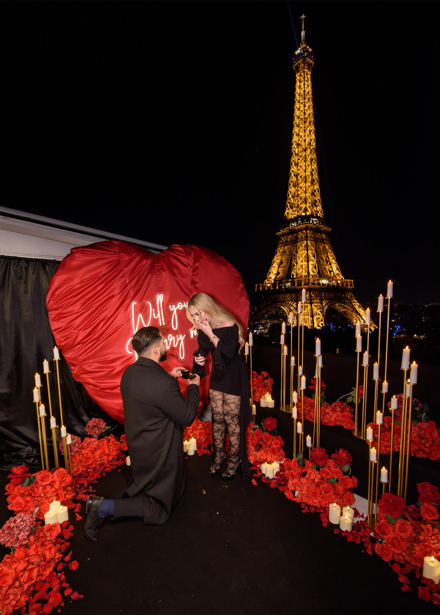 Man on one knee proposing on the Eiffel Terrace rooftop at night, with a red heart backdrop, neon "Will you marry me" sign, red roses and candles, and the illuminated Eiffel Tower behind them