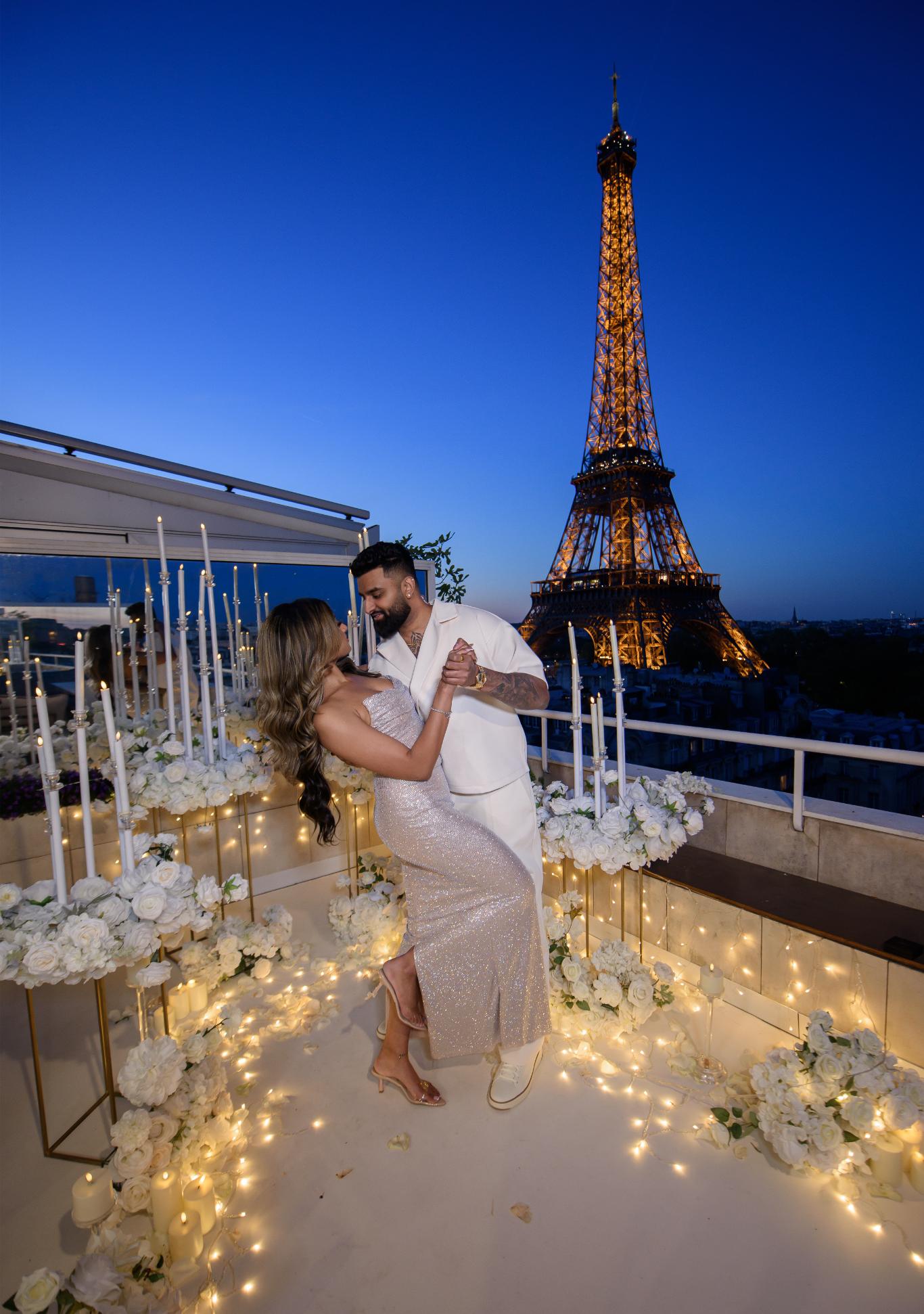 Couple embracing on the Eiffel Terrace rooftop at dusk, surrounded by white floral arrangements, tall candles and fairy lights, with the Eiffel Tower lit up in the background