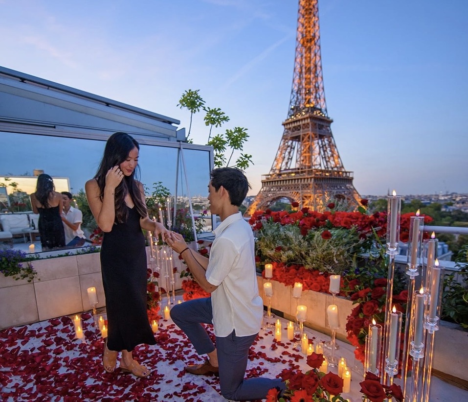 Man on one knee proposing on the Eiffel Terrace rooftop in Paris, surrounded by red roses, rose petals and candles, with the illuminated Eiffel Tower in the background