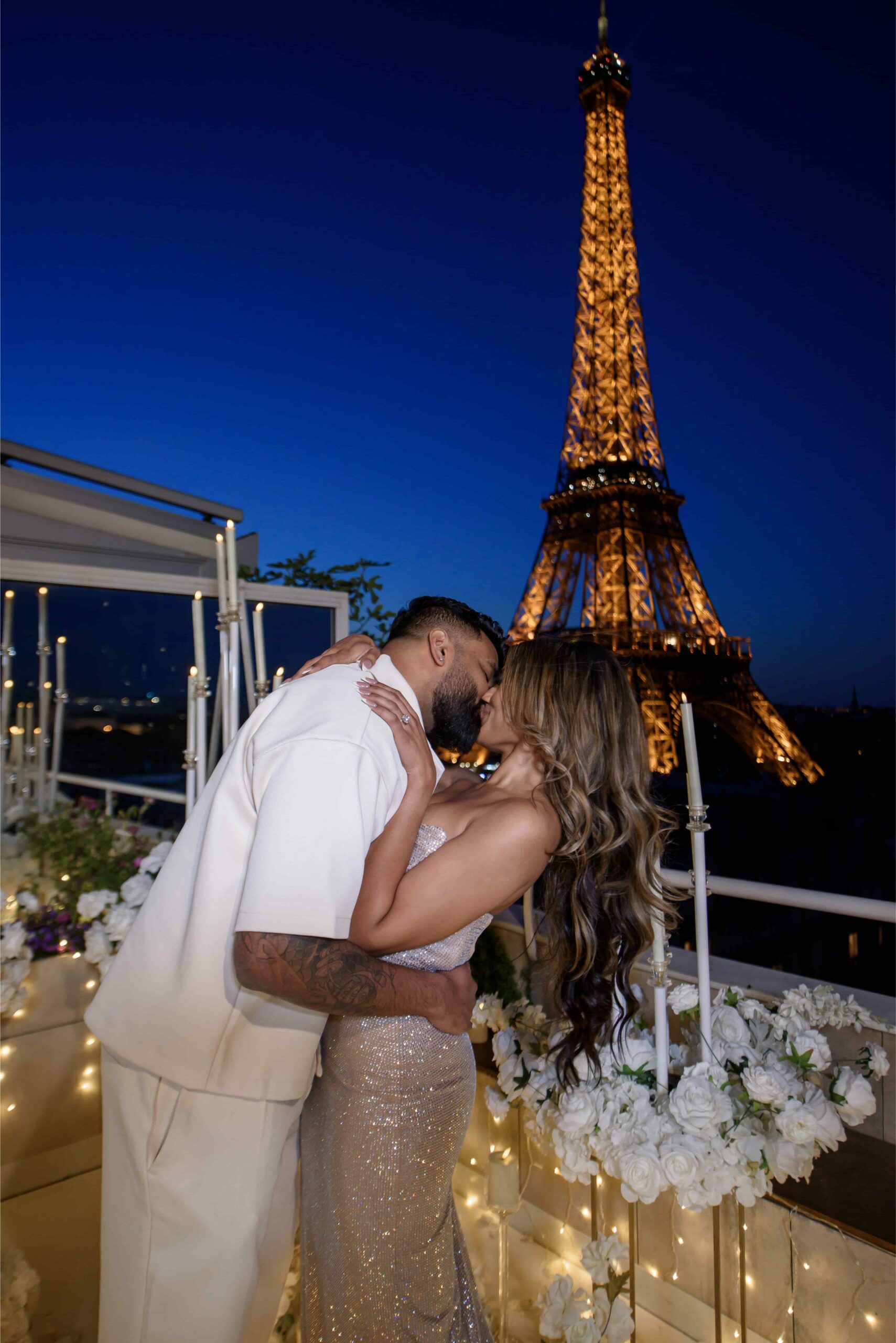 Couple kissing on a candlelit rooftop terrace in Paris, with the illuminated Eiffel Tower glowing against a deep blue night sky, surrounded by white roses and fairy lights