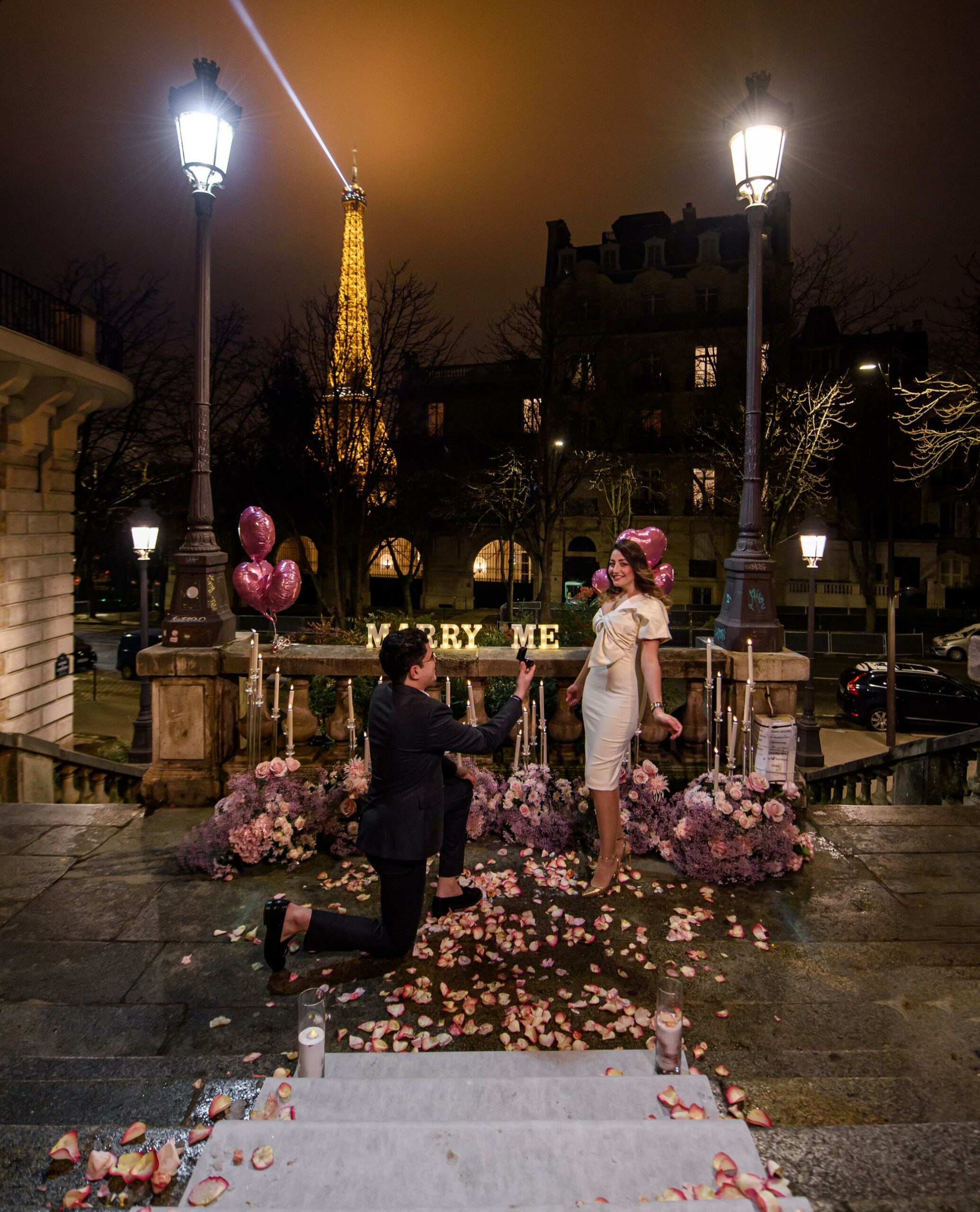 Man proposing on one knee at Avenue de Camoëns in Paris at night, with Marry Me letters, flowers, candles and the illuminated Eiffel Tower in the background