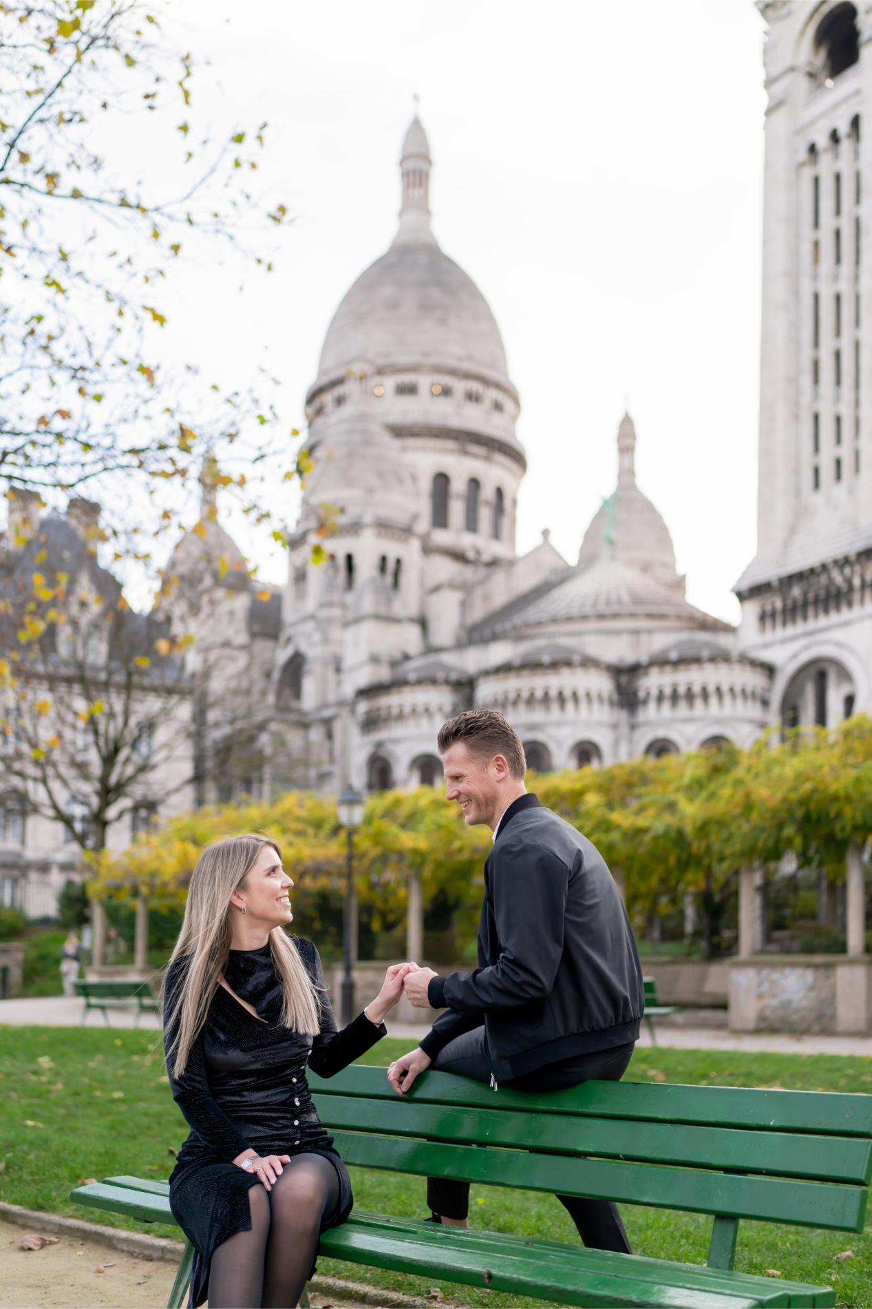 Couple sitting on a green bench in front of Sacré-Coeur basilica in Montmartre, Paris, holding hands and smiling at each other in autumn