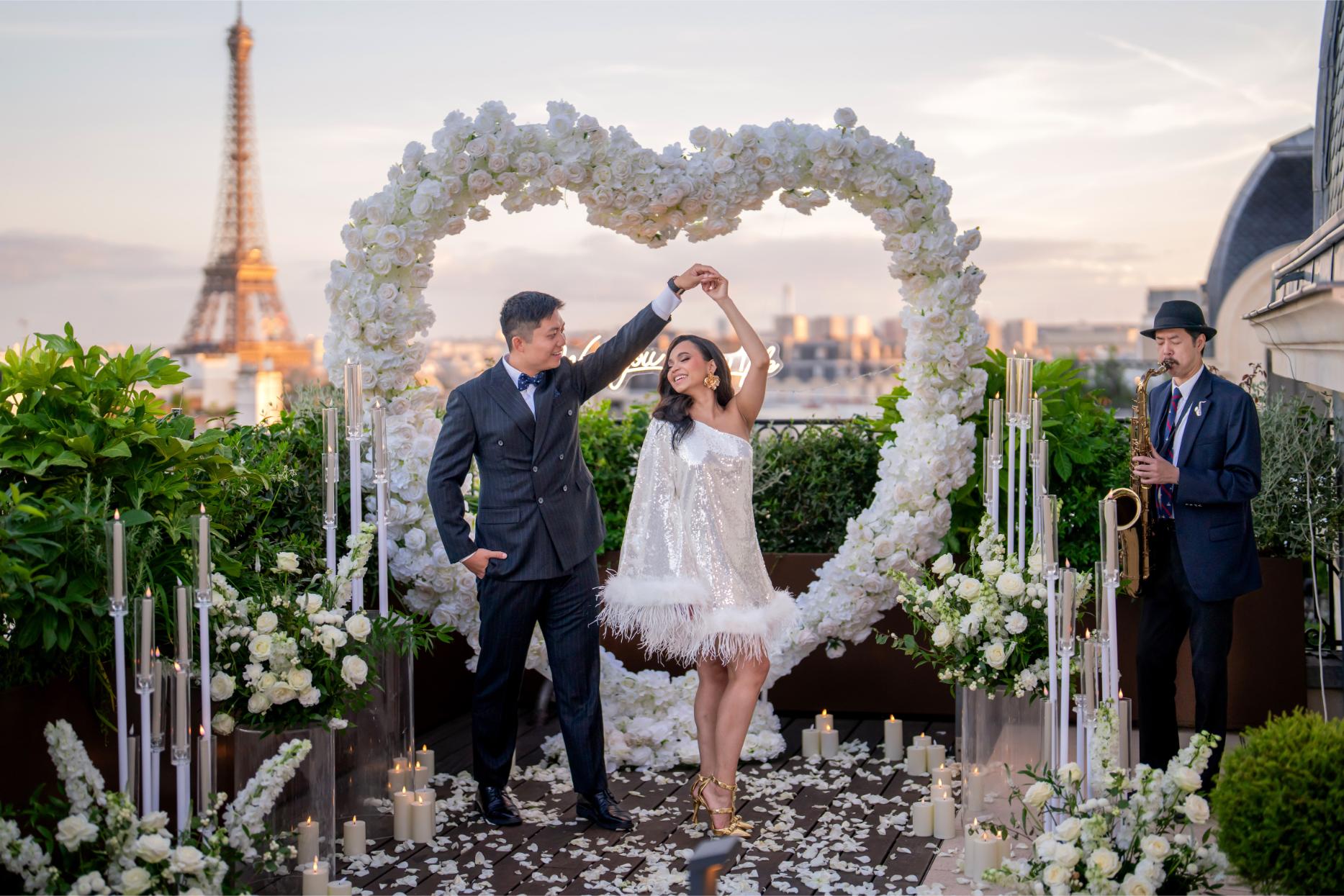 Couple dancing under a white rose heart arch on the Kléber rooftop terrace in Paris, with candles, white floral arrangements, a live saxophonist and the illuminated Eiffel Tower at sunset in the background