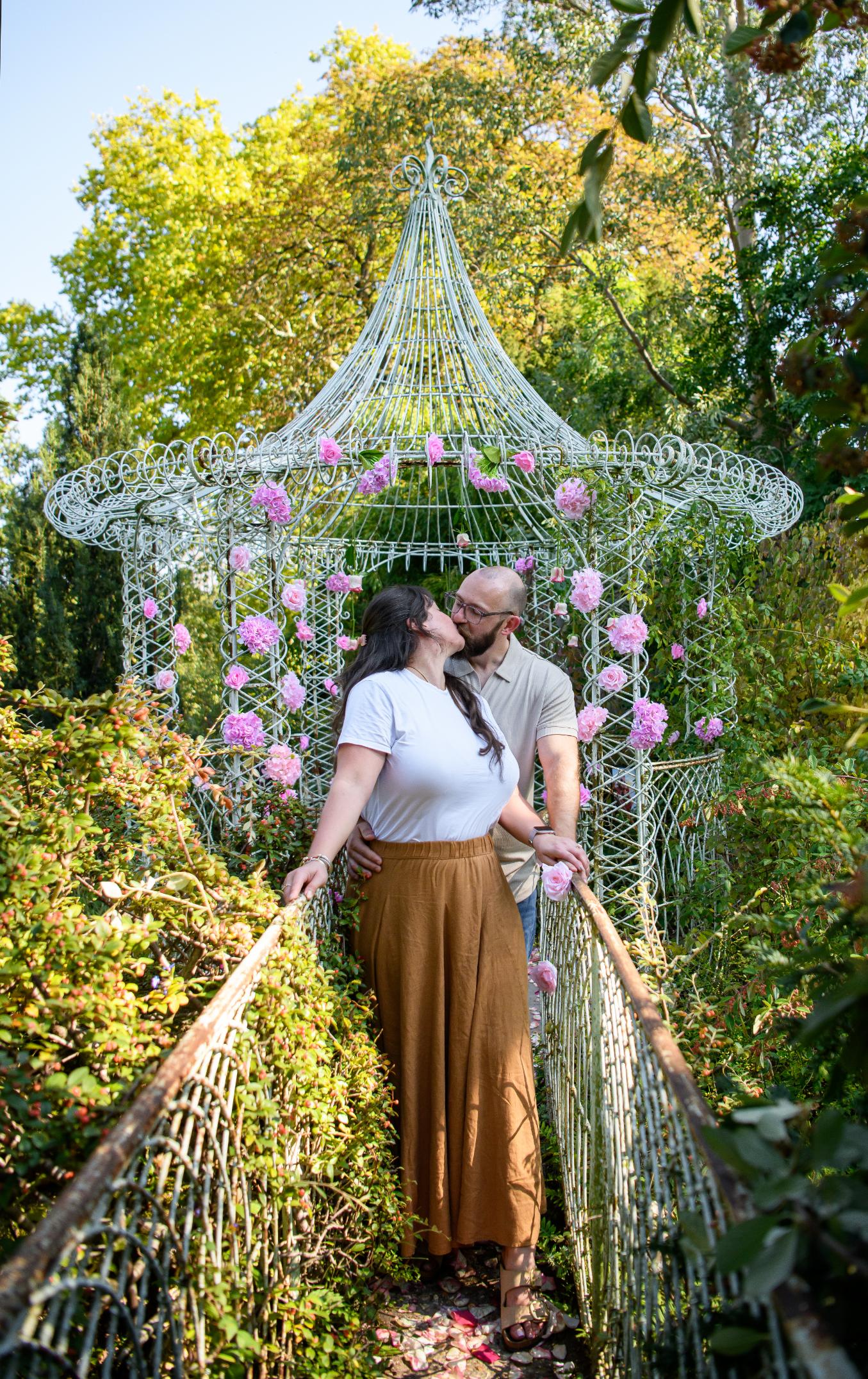 Couple s'embrassant devant une tonnelle en fer forgé ornée de roses roses au Parc de Bagatelle à Paris