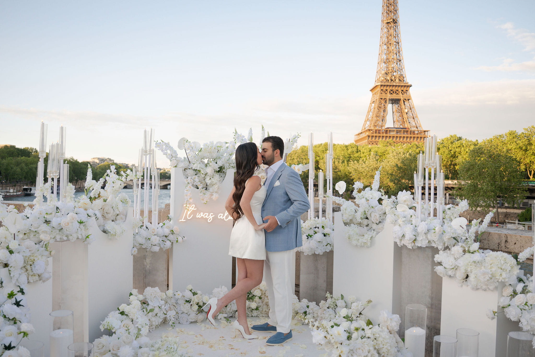 Couple kissing after a marriage proposal in Paris, surrounded by an all-white floral setup with tall candelabras, white flower arrangements and a neon sign, with the Seine River and the Eiffel Tower at sunset in the background