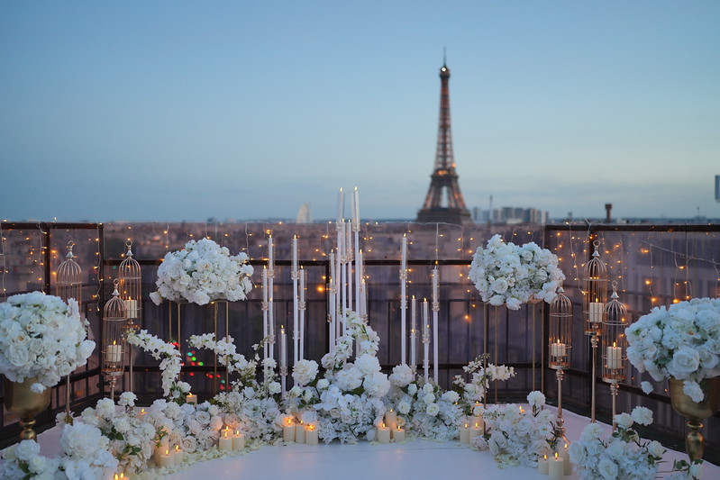 Luxury proposal setup on a private rooftop terrace in Paris at dusk, with white flower arrangements, tall candles, decorative bird cages and fairy lights, and the Eiffel Tower in the background