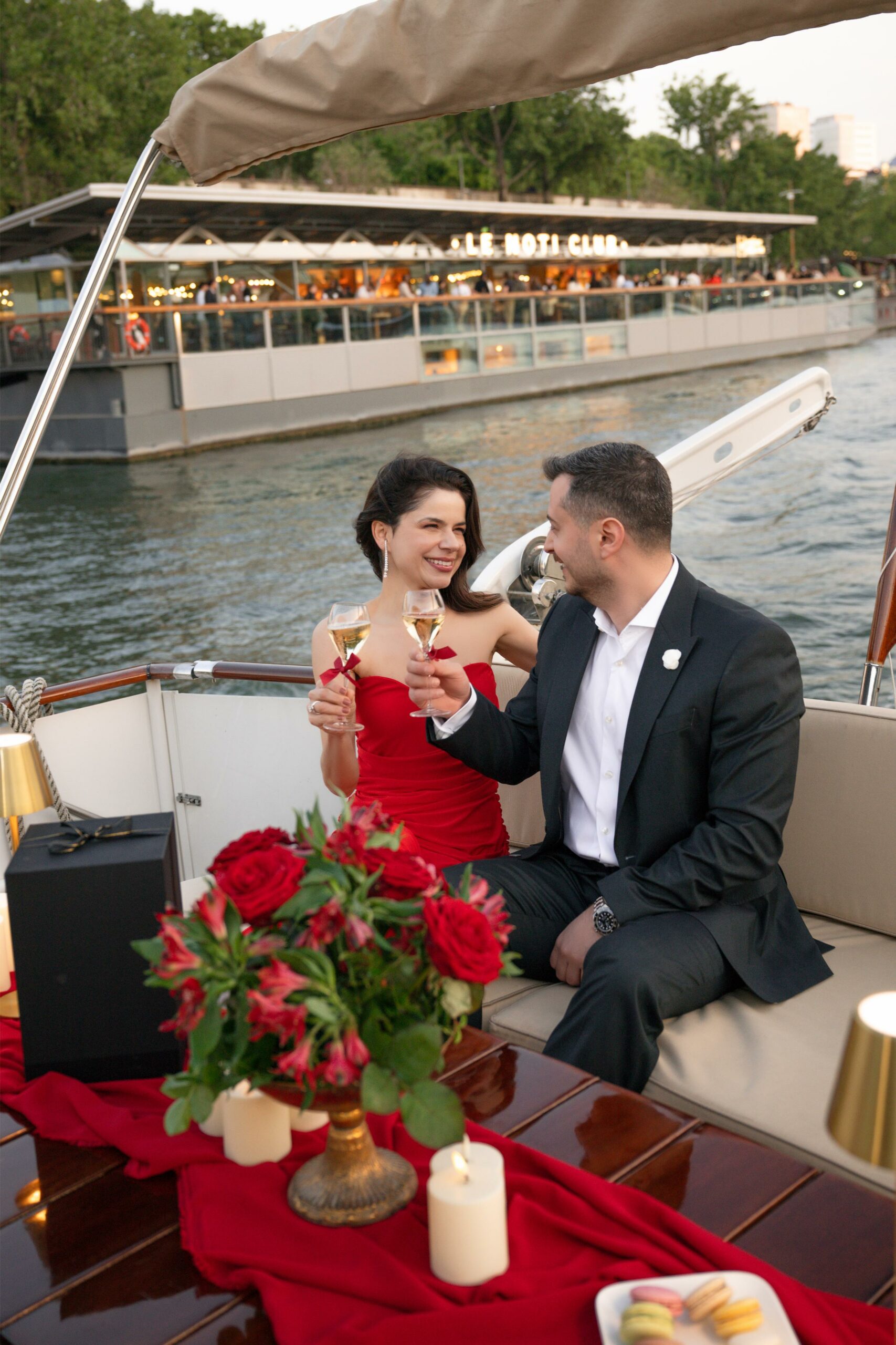 Couple toasting with champagne on a private boat cruise on the Seine in Paris, with red roses, candles and macarons on the table at sunset